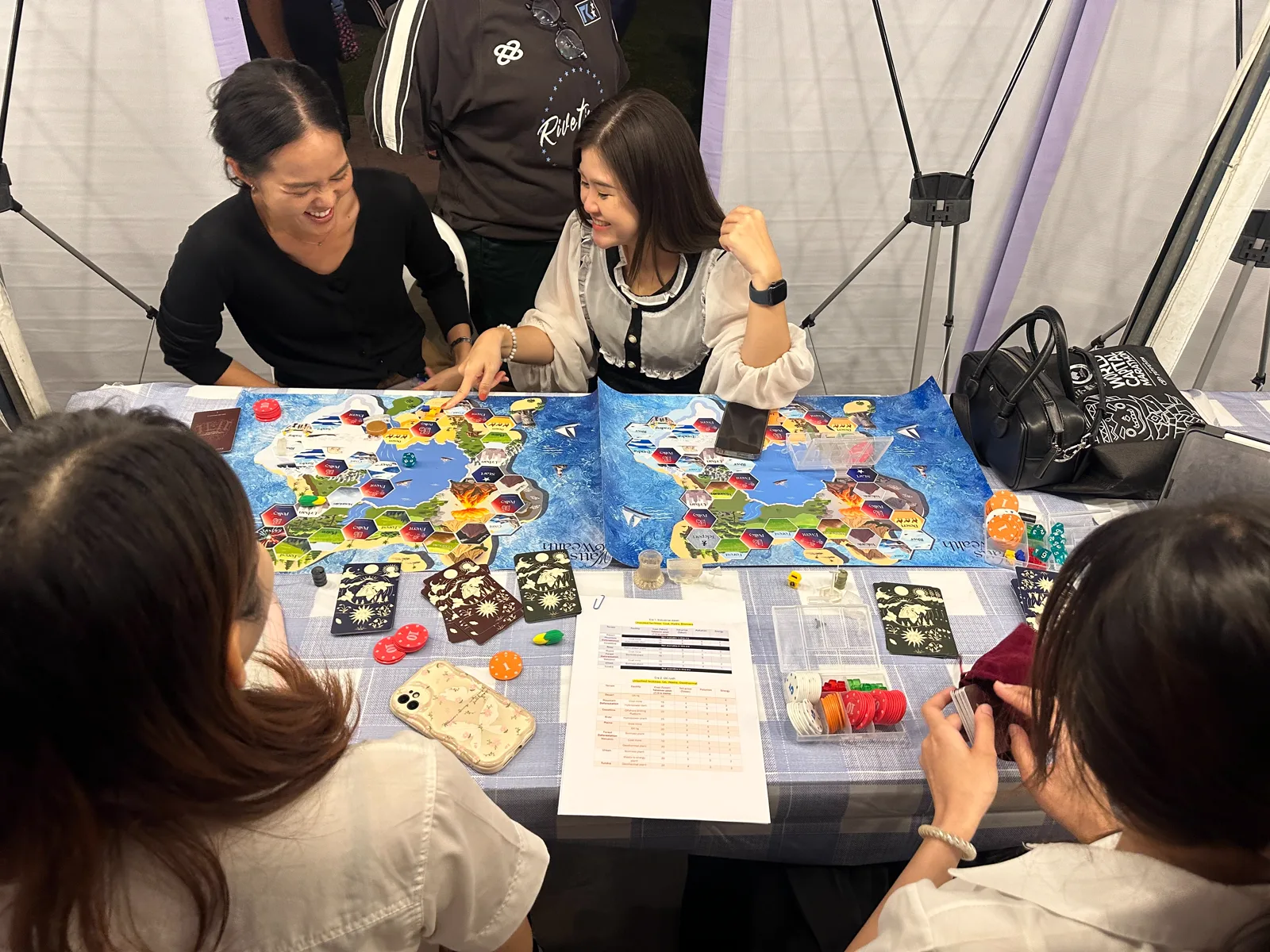 Players around two physical Watts and Wealth boards mid-session — hex-tile world maps, card decks, dice, and token chips on the table